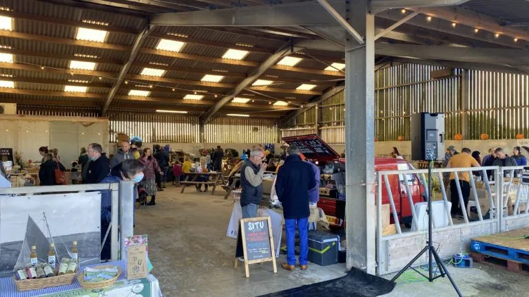 A traditional Cornish farm shop barn with stalls of local vegetables, meats, and cheeses, customers browsing the produce