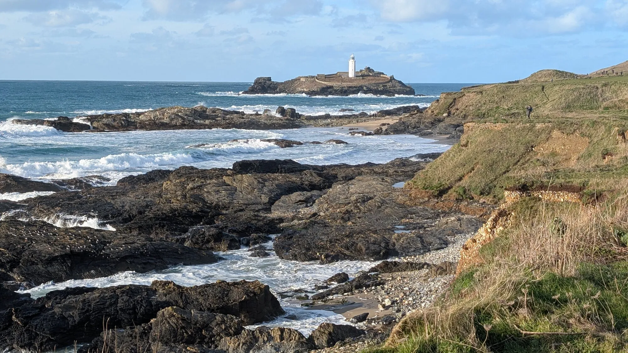 Godrevy Lighthouse on its rocky island off Cornwall's north coast, viewed from the rugged South West Coast Path with waves breaking over dark rocks in the foreground.