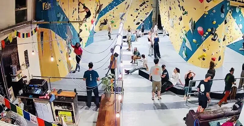 Climbers on the colourful angled bouldering walls inside BoulderRyn in Penryn, with thick crash matting below and a relaxed crowd watching.