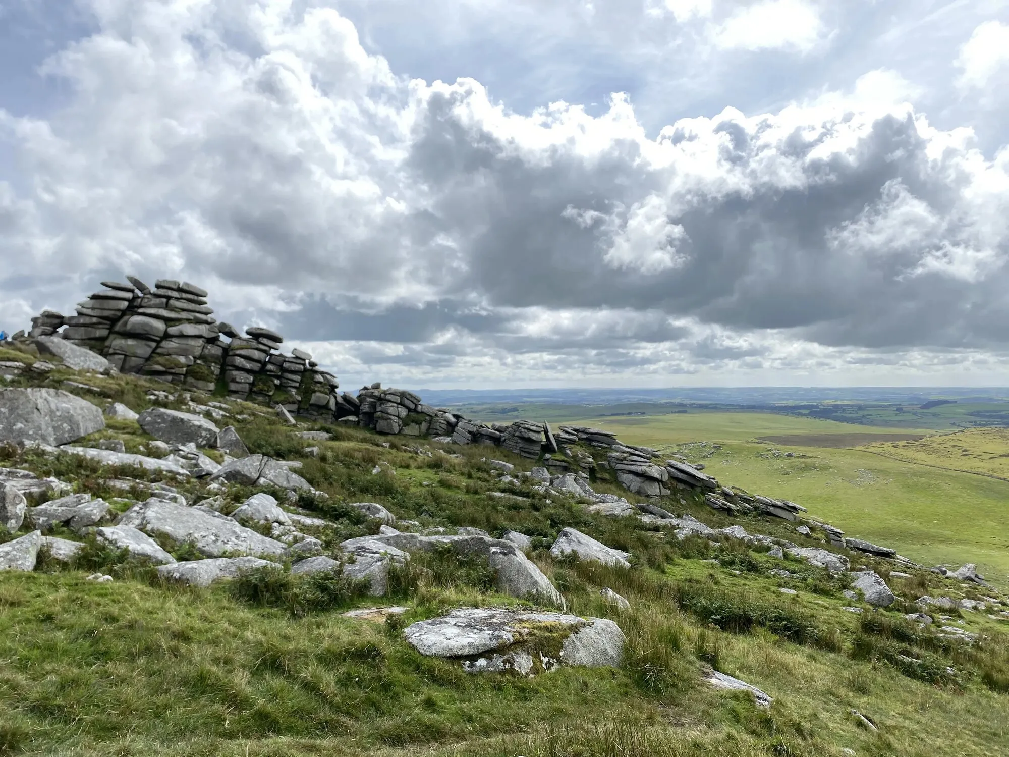 The summit of Rough Tor on Bodmin Moor, with stacked weathered granite slabs forming a natural rampart along the ridge, scattered boulders and rough grass in the foreground, and the open moorland stretching out to a wide cloudy sky beyond.