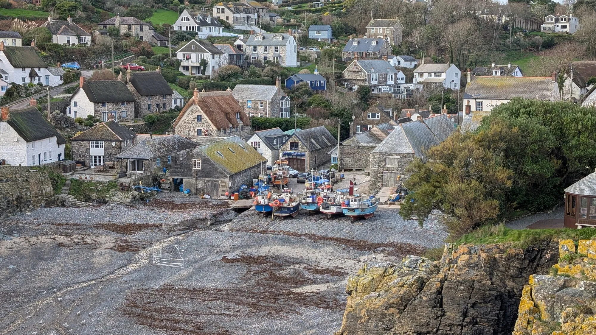 Cadgwith Cove on the eastern Lizard Peninsula in Cornwall, with thatched-roofed fisherman's cottages clustered at the back of a pebble beach, fishing boats hauled up on the slipway, and the small cove sheltered by cliffs.