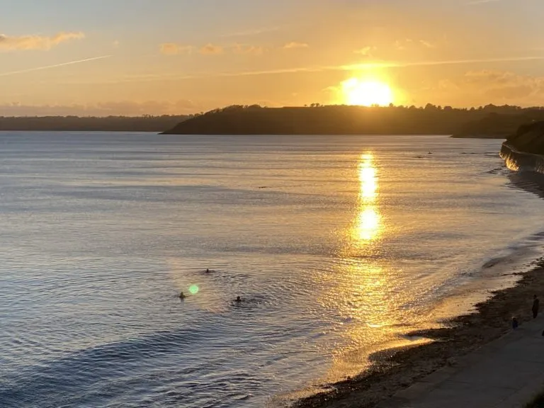 Sunrise over Falmouth Bay seen from above Castle Beach, with two swimmers in the calm water and the path along the wall in the foreground.