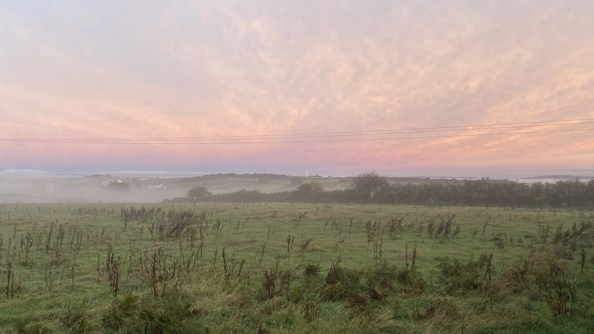 A soft pink dawn over misty fields on the rural edge of Falmouth near Trewena, with low mist lying in the valley and rough meadow grass in the foreground, the kind of quiet Cornish morning the old beliefs were made in.
