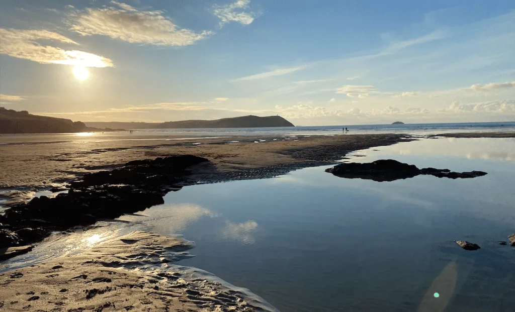 Low tide on a Cornish beach at golden hour, sun setting behind a distant headland, with wet sand reflecting the sky and a calm tidepool in the foreground.