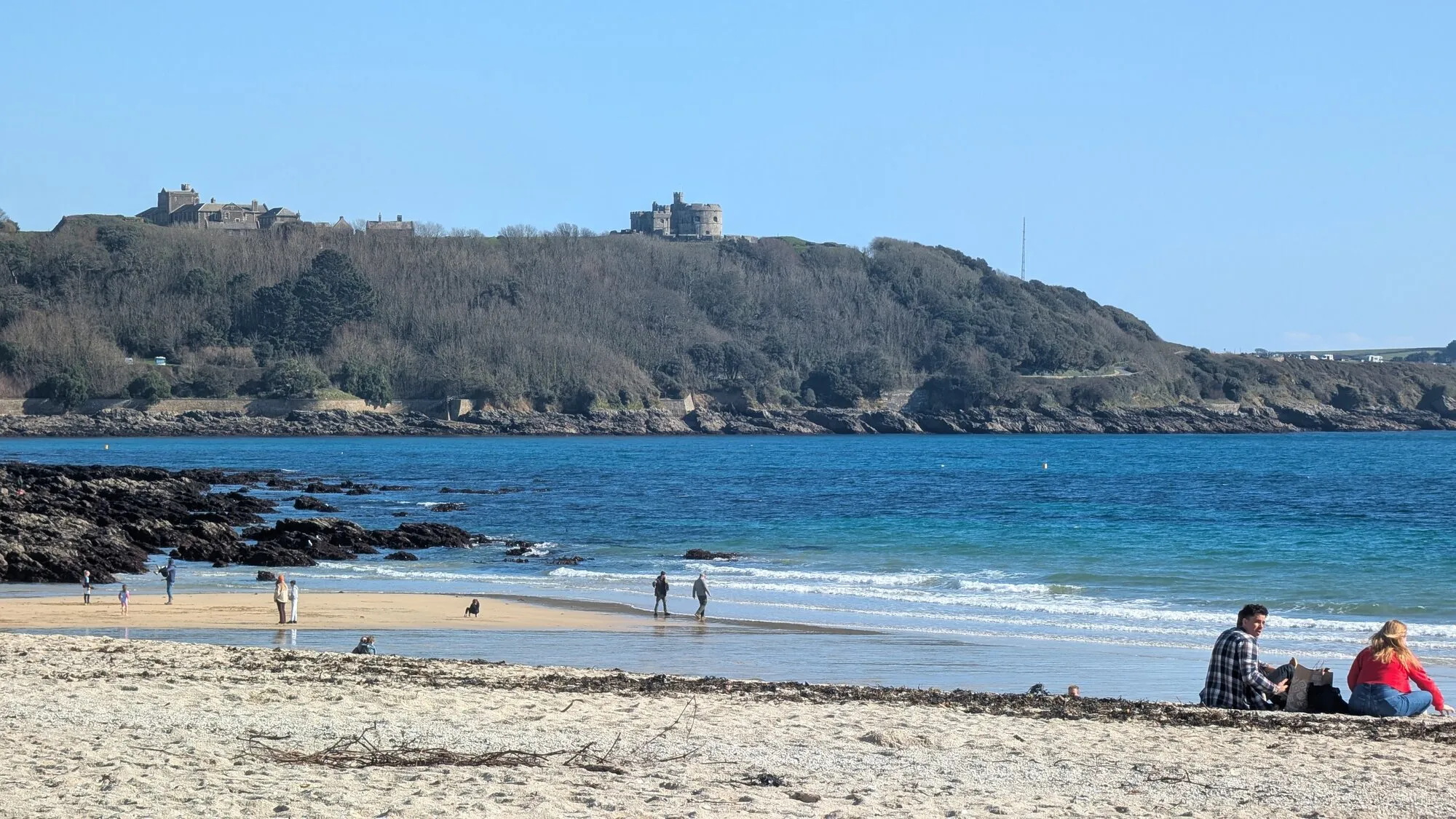 A couple sitting together on the sand at Gyllyngvase Beach in Falmouth on a clear spring day, with Pendennis Castle visible on the wooded headland in the background, the rocks of the eastern end of the beach to the left, gentle waves lapping the shoreline and a few other beachgoers walking along the wet sand.