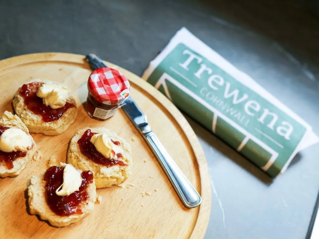 Cornish scones served jam first then cream, on a wooden board beside a Trewena Cornwall leaflet, with a small jar of strawberry jam, a knife and a butter-curl on each scone.