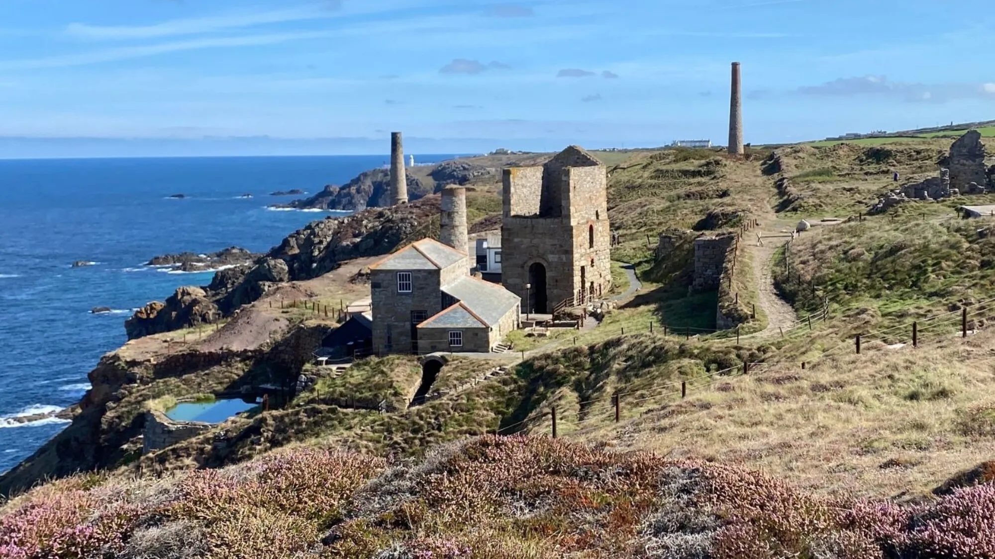 Ruined tin-mine engine houses and chimney stacks standing on the cliffs of Cornwall's Tin Coast near St Just, above a bright blue Atlantic sea with heather in the foreground, part of the Cornish Mining World Heritage landscape.