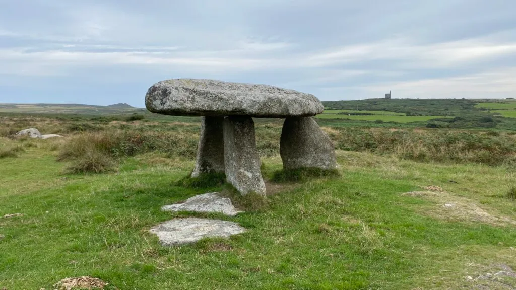 Lanyon Quoit, a Neolithic dolmen on the Penwith Moors. A massive granite capstone resting on three upright slabs, with green fields and an old mining chimney on the horizon.