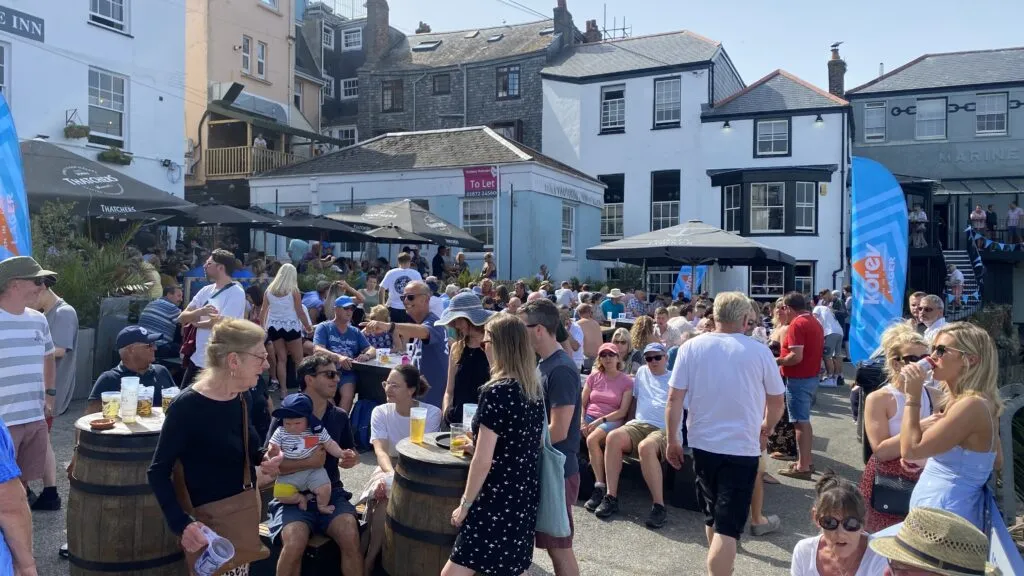 A busy summer afternoon on the quayside outside the Chain Locker pub in Falmouth, with a large crowd drinking and chatting around upturned-barrel tables on a sunny event day.