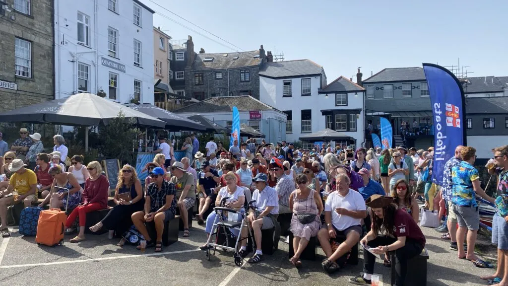 A large crowd watching a sea shanty performance on Custom House Quay in Falmouth on a sunny day, with the Quayside Inn pub in the background and RNLI Lifeboats branding banners visible alongside the audience.