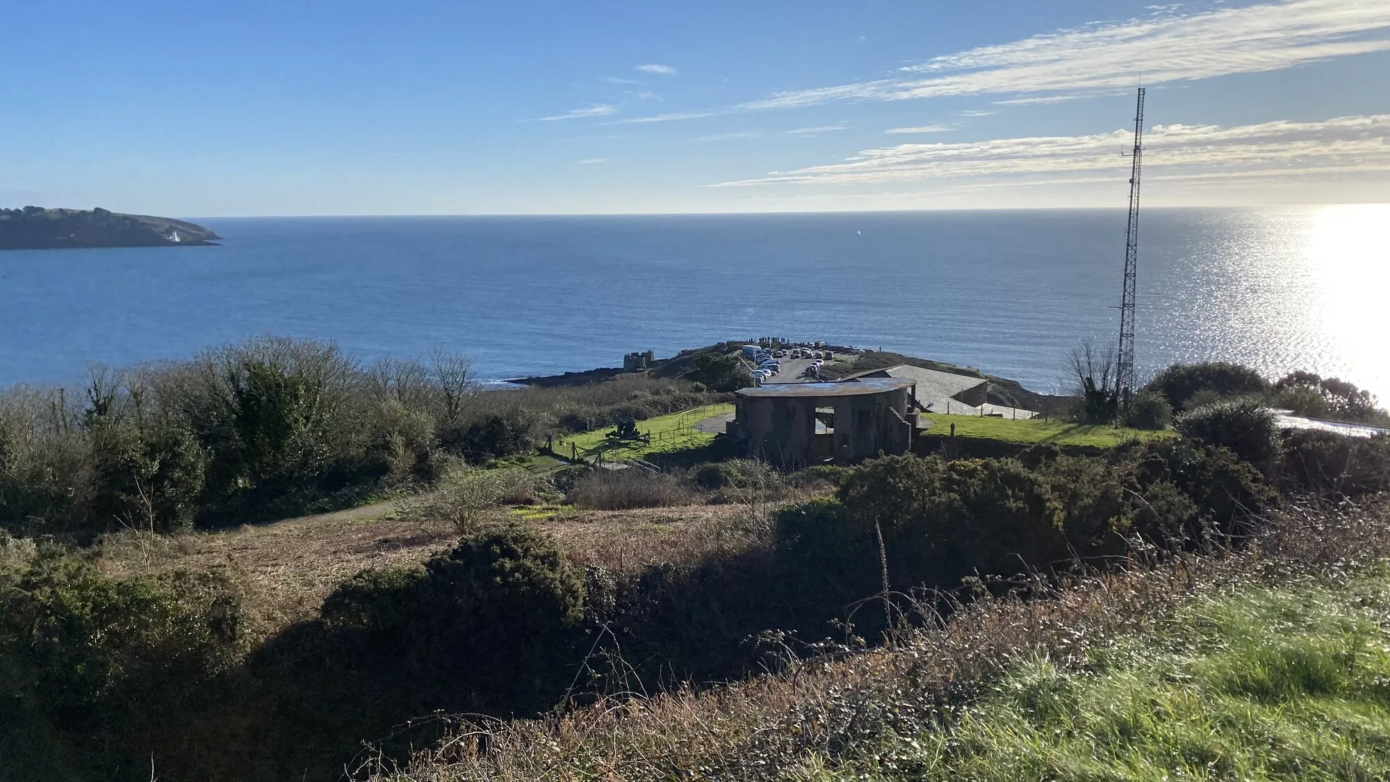 Looking out from Pendennis Point in Falmouth on a clear winter day: a preserved WWII concrete gun emplacement and tall aerial mast stand on the open headland, with calm blue water stretching to the horizon, the white lighthouse on St Anthony's Head visible on the far headland to the left, and dry grasses and gorse in the foreground.