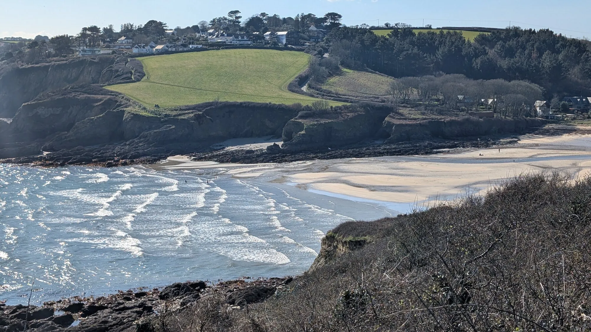 Maenporth beach viewed from the coast path above Gylly, with white surf rolling onto a wide curve of low-tide sand, green cliff-top fields and village houses on the headland beyond.
