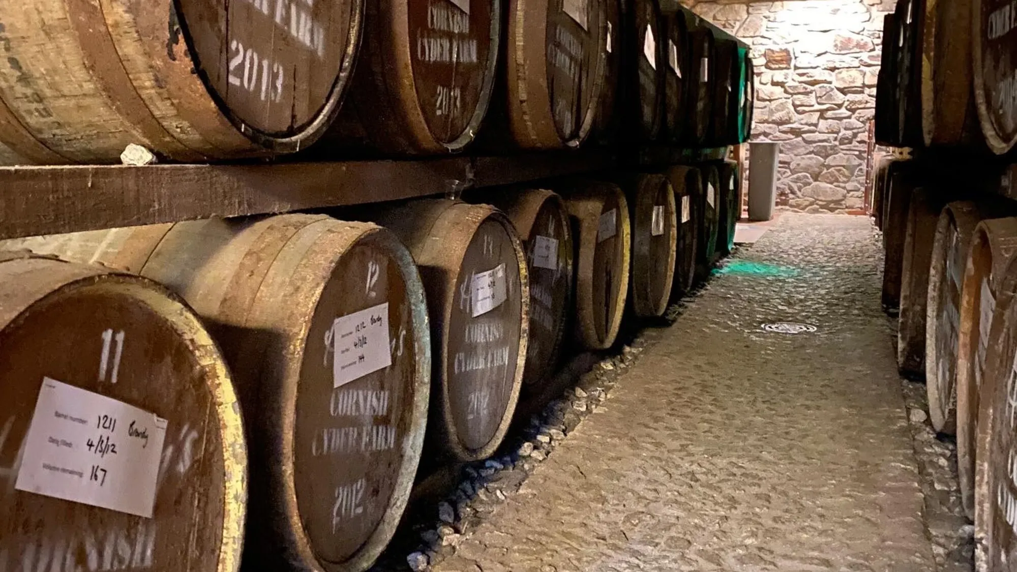 Rows of oak cyder barrels in the dimly lit cellar at Healey's Cornish Cyder Farm near Truro, with the Cornish Cyder Farm logo painted on the barrel ends, dates marked from 2012 and 2013, and the cobbled stone floor of the maturation room.