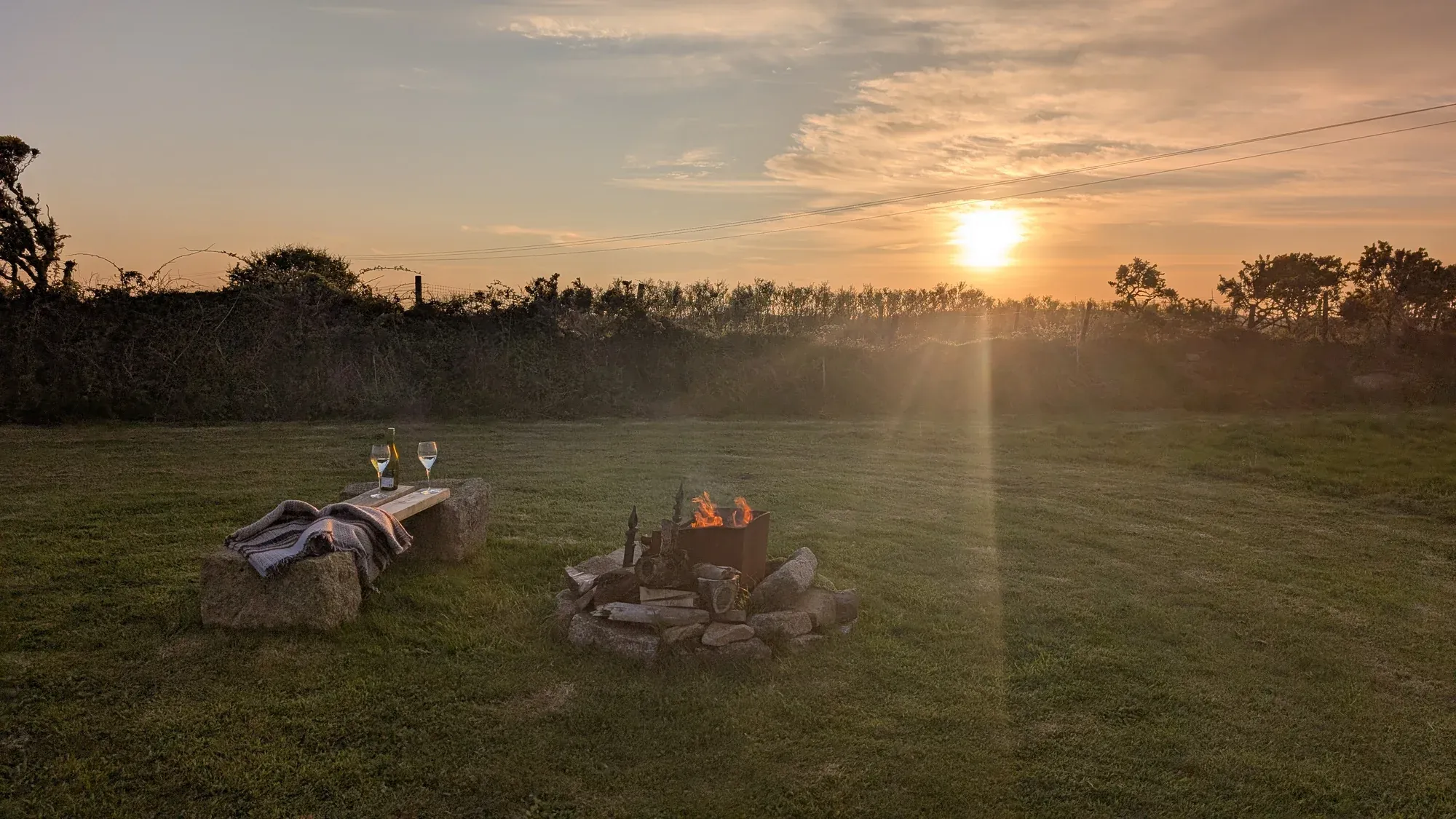 Sunset over the fields at Trewena Cottages, with a small fire burning in a stone fire pit, a granite bench beside it set with two glasses of white wine, a soft grey blanket draped across the bench, and the Cornish horizon glowing orange behind.