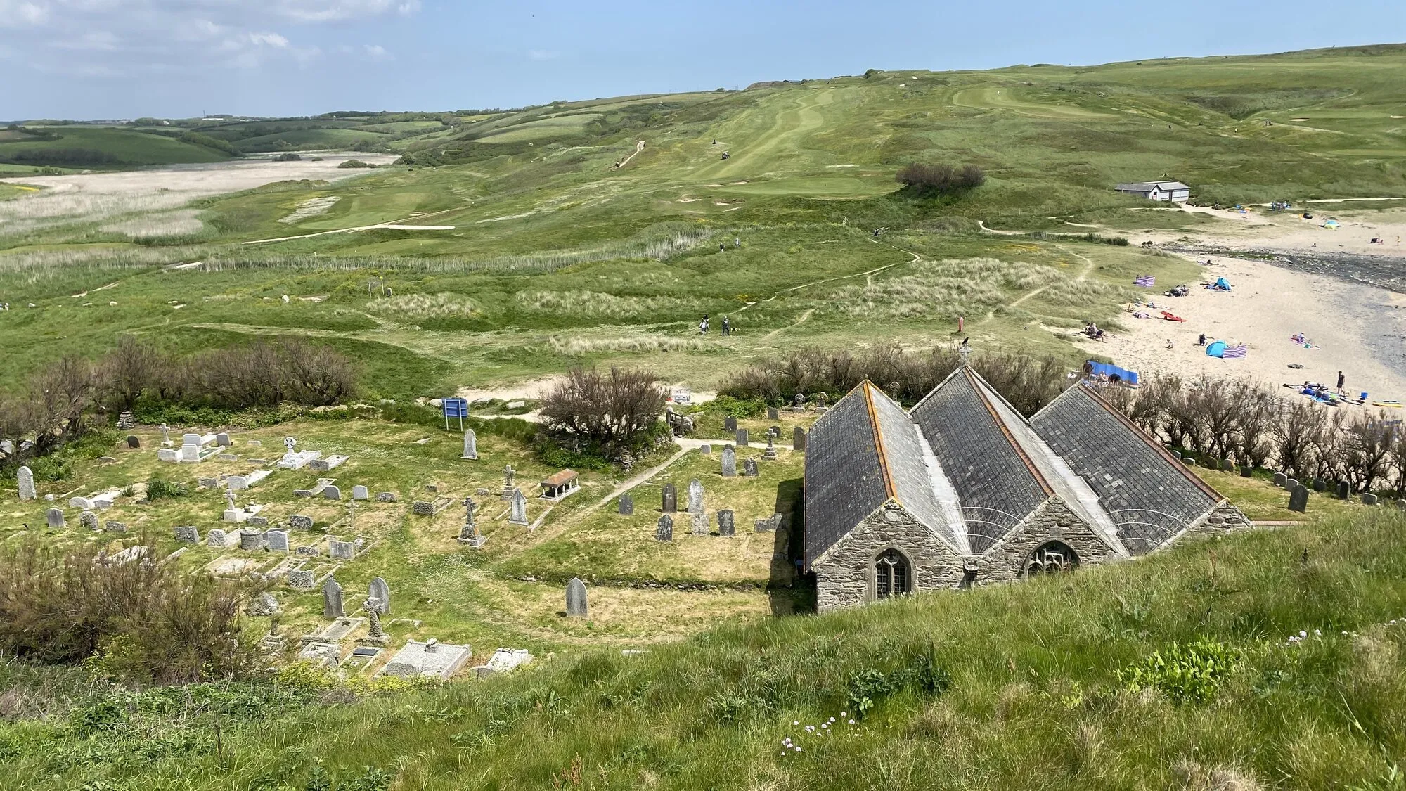 Church Cove at Gunwalloe on Cornwall's Lizard Peninsula, with the 14th-century Church of St Winwallow built right onto the edge of the beach, an ancient graveyard with weathered Celtic crosses, and a wide sandy beach with families on it leading off to the right.