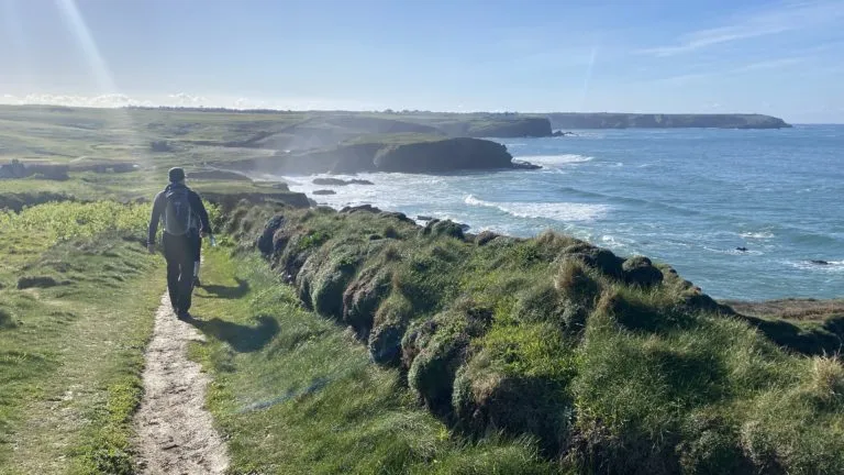 A hiker walking along a clifftop section of the South West Coast Path on the Lizard Peninsula, with dramatic curving coastline and the Atlantic stretching into the distance.