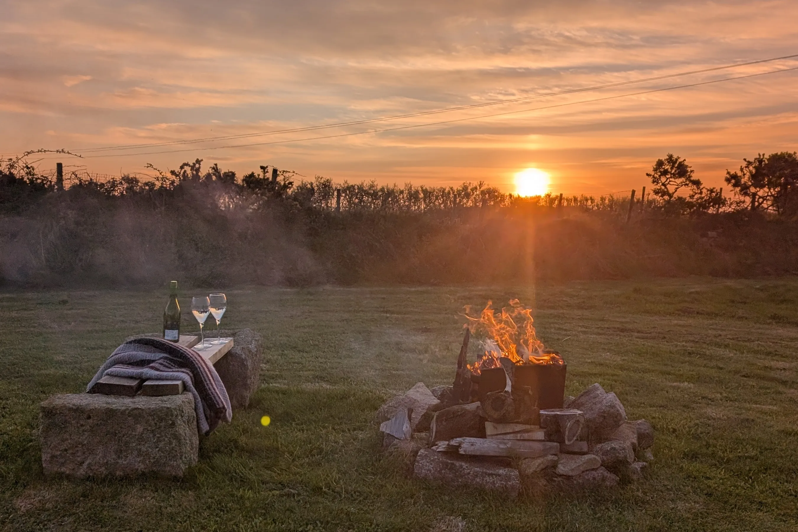 The fire pit in the meadow at sunset, a bench with wine and glasses to one side, sky burning orange.