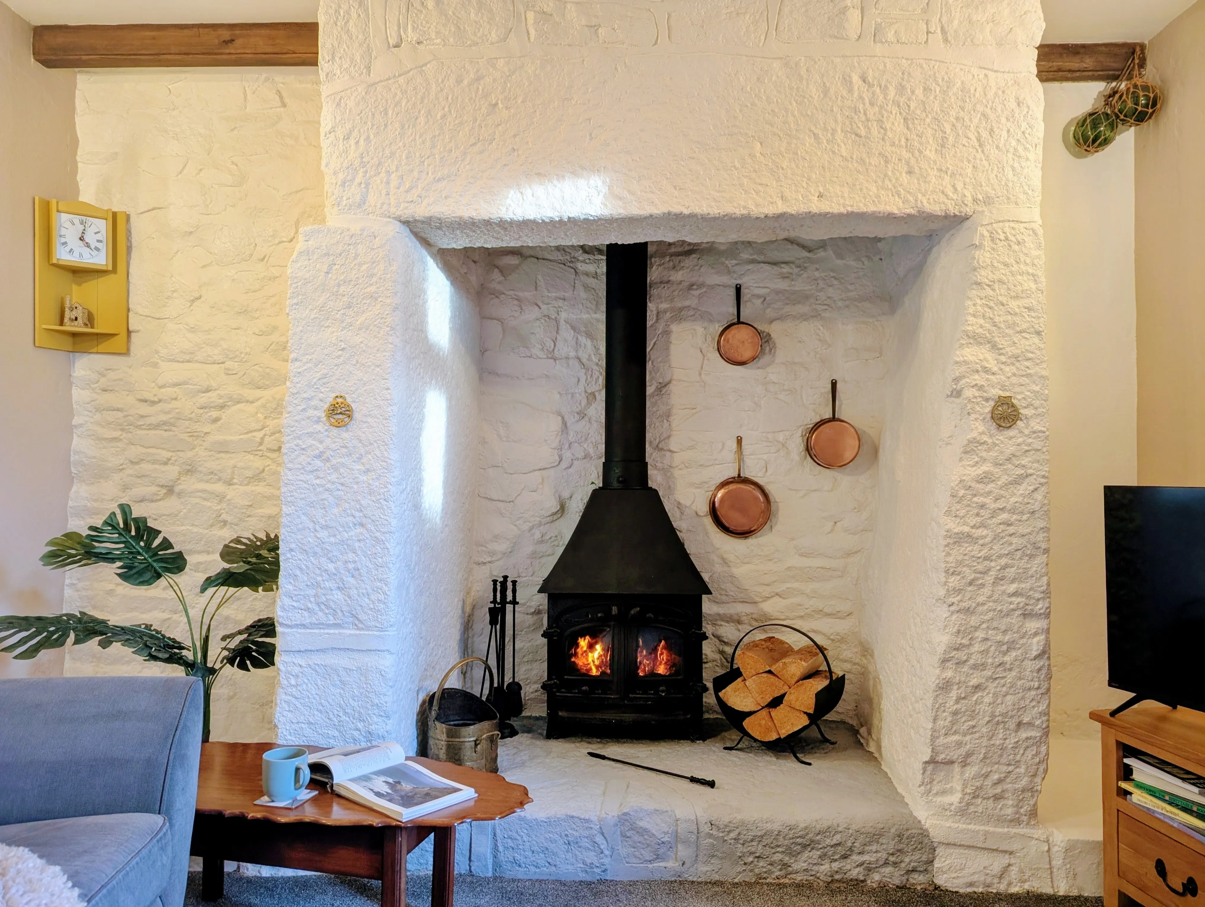 The wood burner in Little Avalon's granite fireplace, copper pans hanging on the white wall, log basket beside.