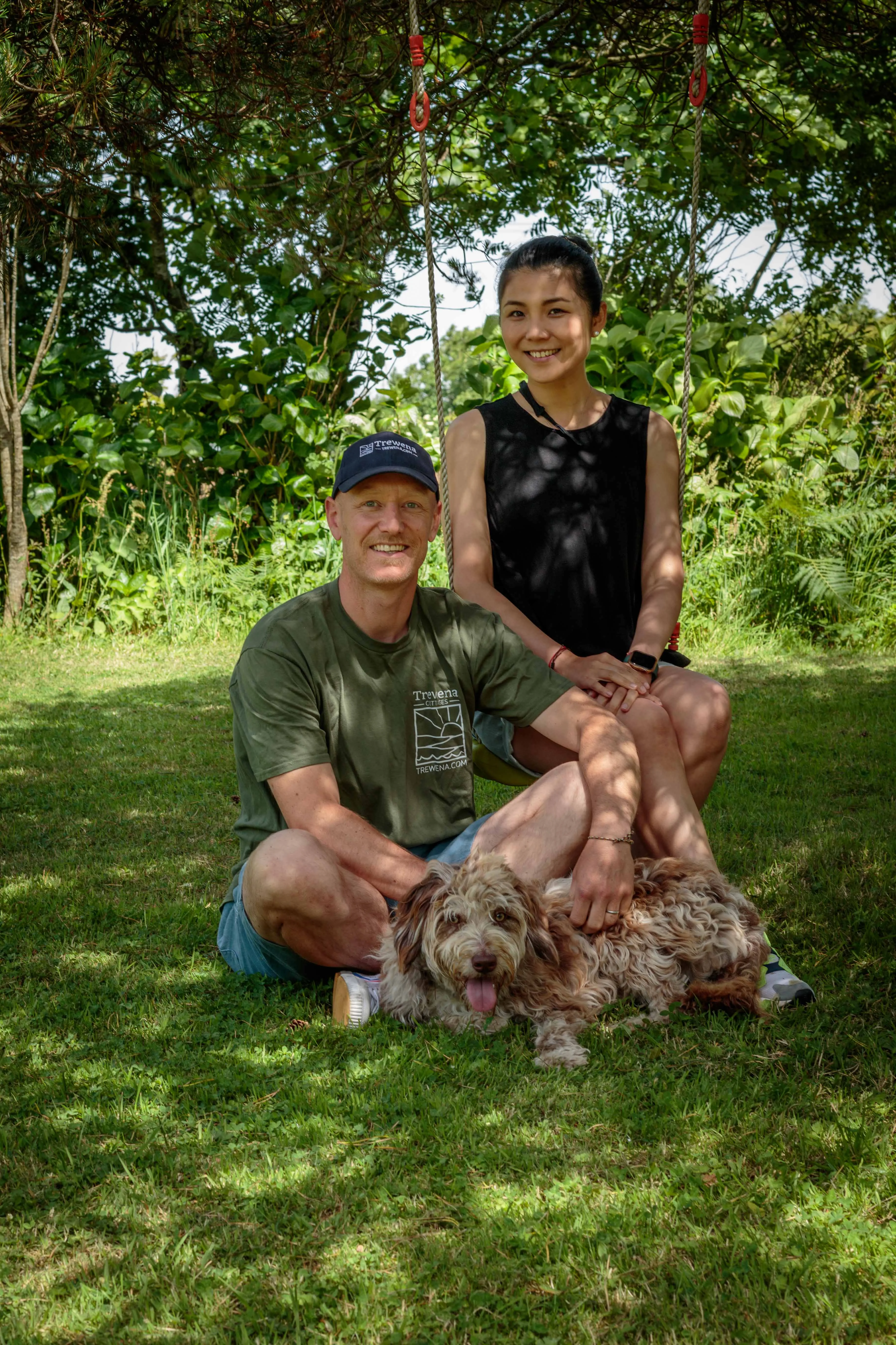 Rich sitting cross-legged with the dog on his lap, Hellen on a swing behind, in the dappled light of the woods at Trewena.
