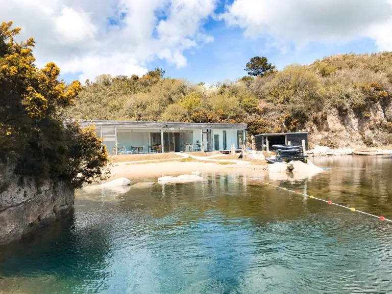 The visitor centre at Kernow Adventure Park sits on a small sandy beach beside the flooded waters of Kessel Quarry, with granite slopes covered in yellow gorse rising behind it and a swim-lane line on the calm water in the foreground.
