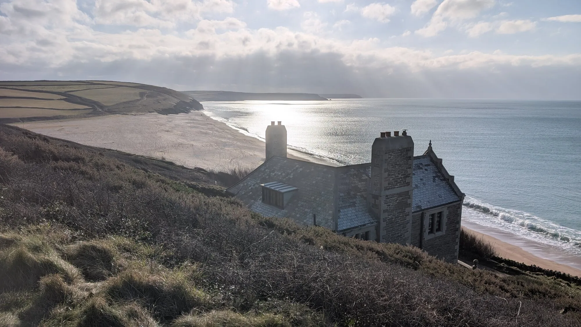 A slate-roofed stone house on the slope above Loe Bar, with the long sweep of the bar below, sunlight shining on the sea and the coast running away towards the Lizard.