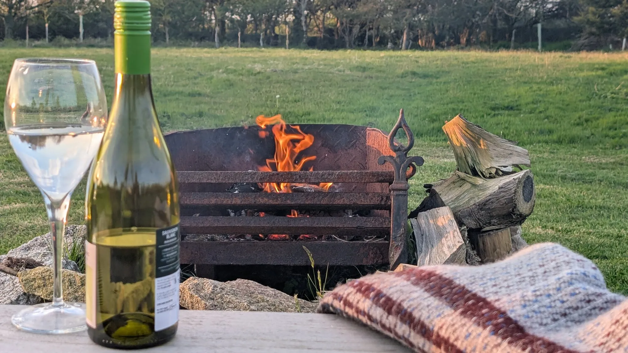 A bottle of white wine and a tall glass on a wooden table beside a lit firepit in the late-afternoon sun, with a wool throw over the corner of the table and a green meadow stretching away into golden Cornish trees behind.