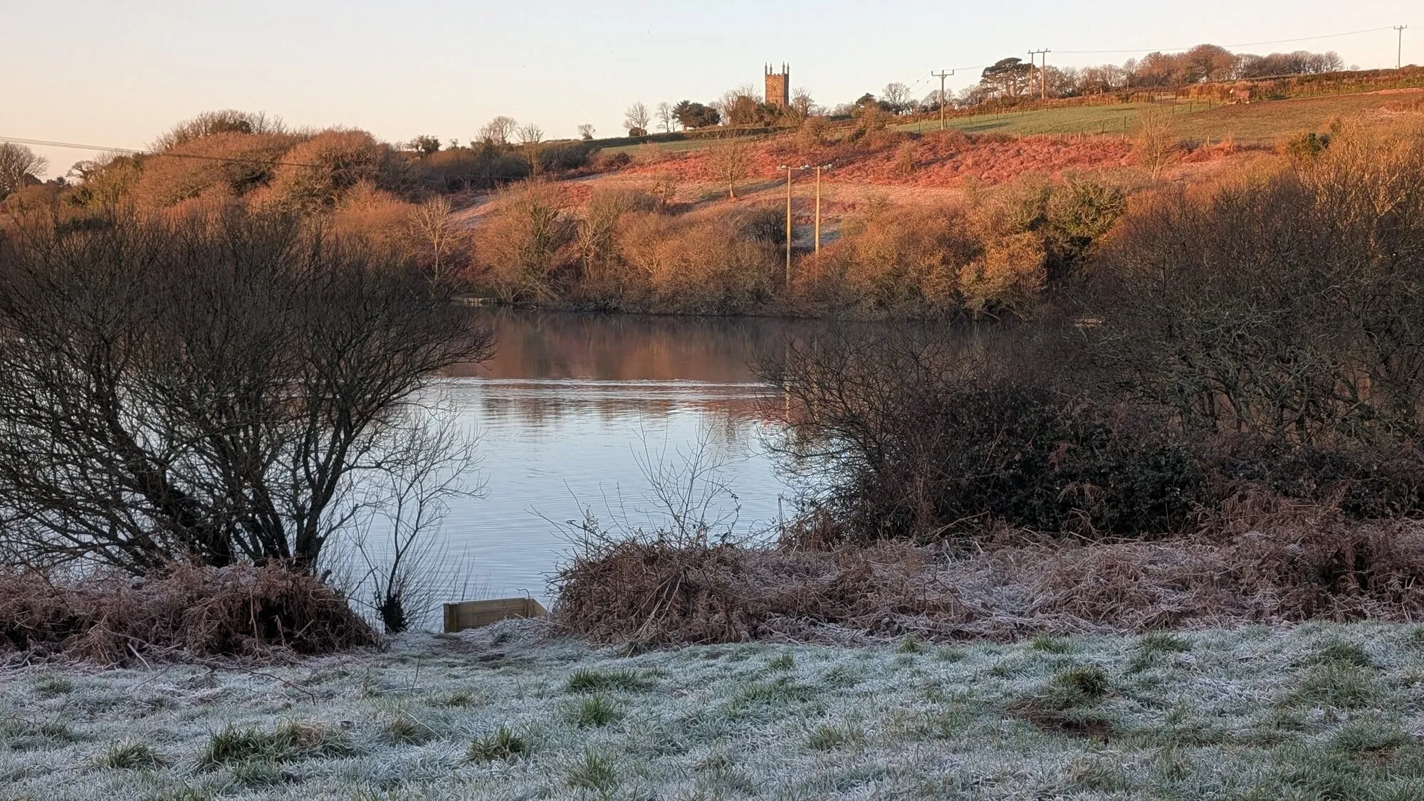 Argal Reservoir in Mabe parish on a frosty winter dawn: the still water of the reservoir reflecting the rising sun's golden light, with bare-branched trees and bracken on the far slope, the medieval tower of St Laudus Church visible on the hilltop above the wooded shore, and frosted grass in the foreground.
