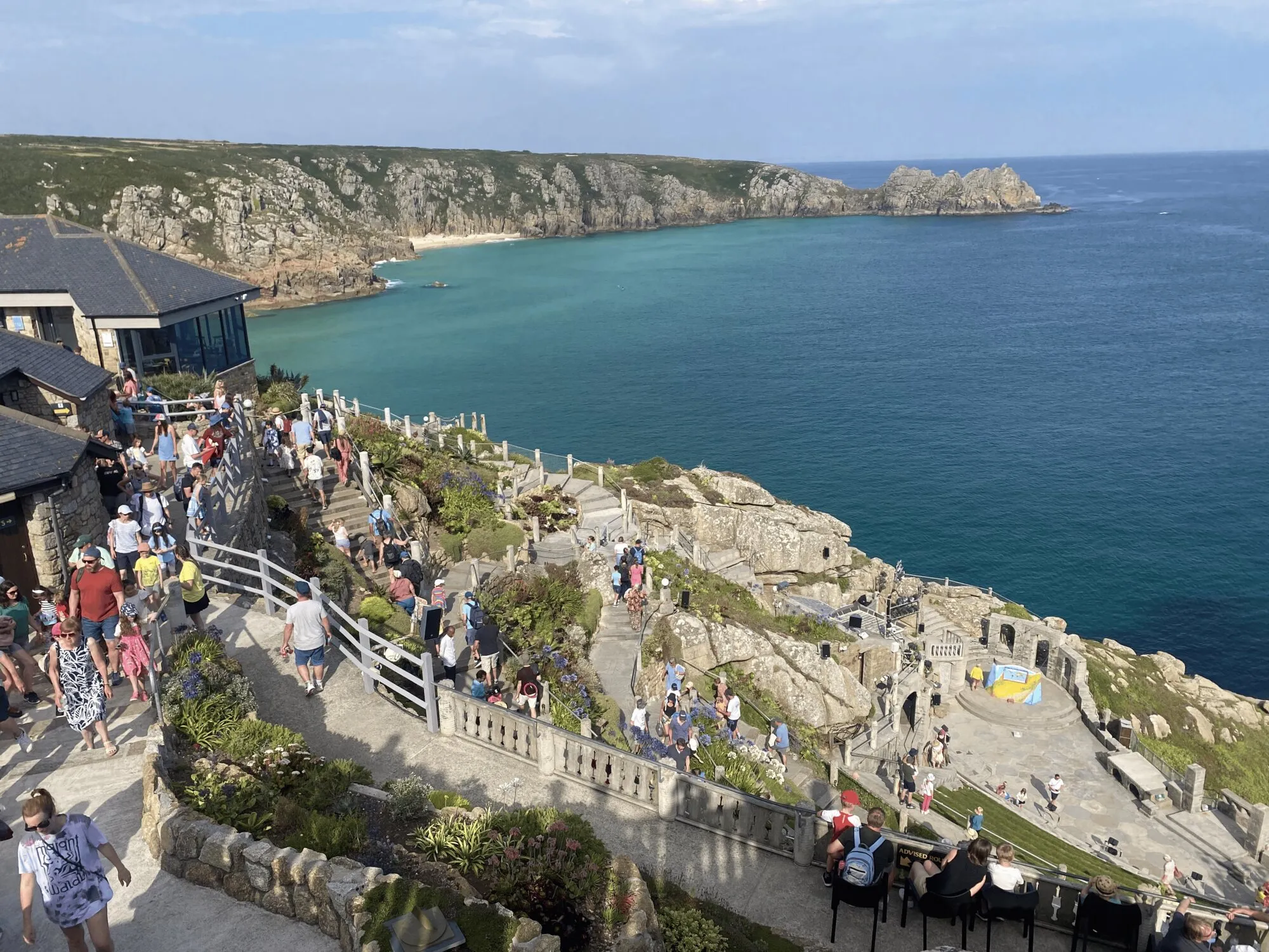 The Minack Theatre at Porthcurno on Cornwall's far western coast, with the granite-cut amphitheatre carved into the cliffside descending towards a turquoise bay, Logan Rock visible in the distance and a clear summer sky overhead.