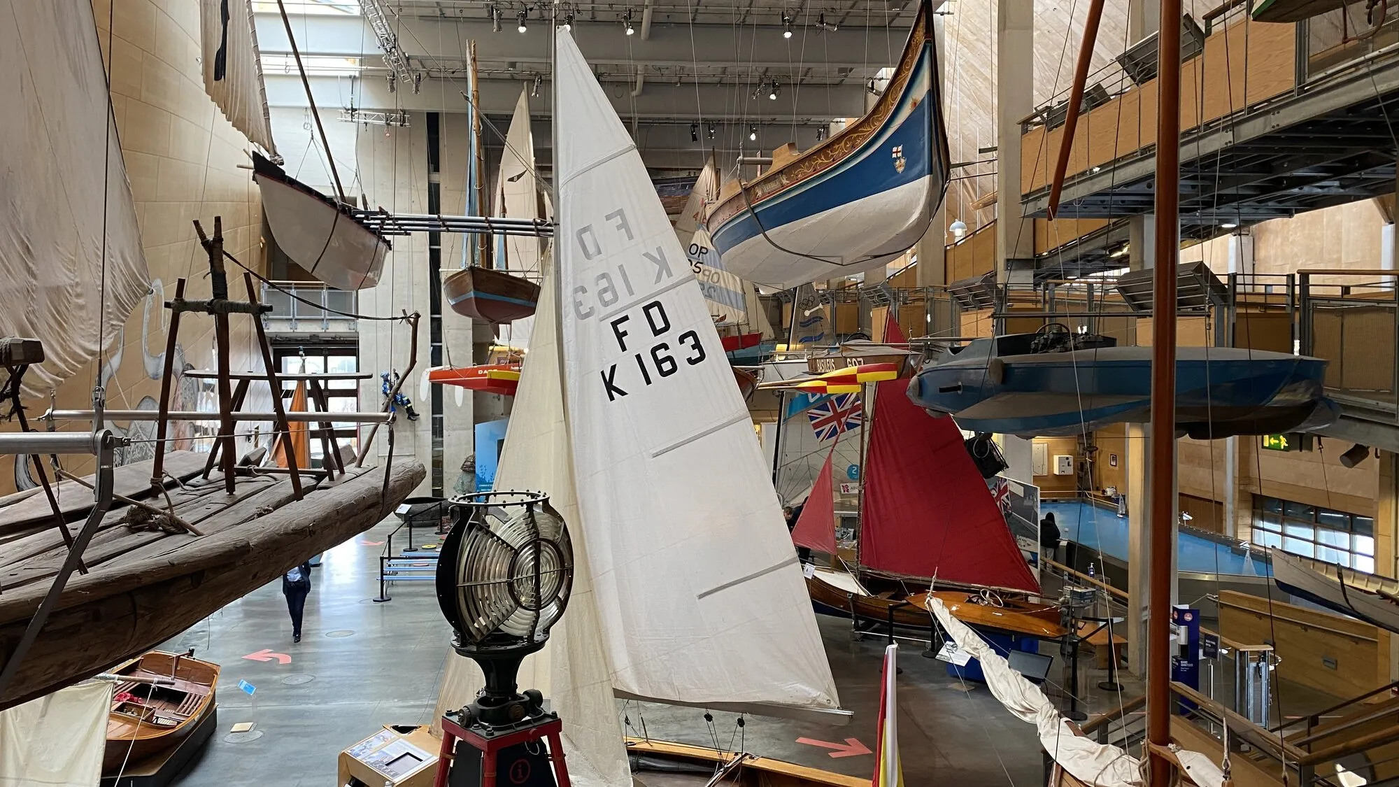 The cavernous main hall of the National Maritime Museum Cornwall in Falmouth, with traditional sailing dinghies, racing yachts and small craft suspended at multiple heights from the ceiling, sails extended, viewed from one of the upper galleries.