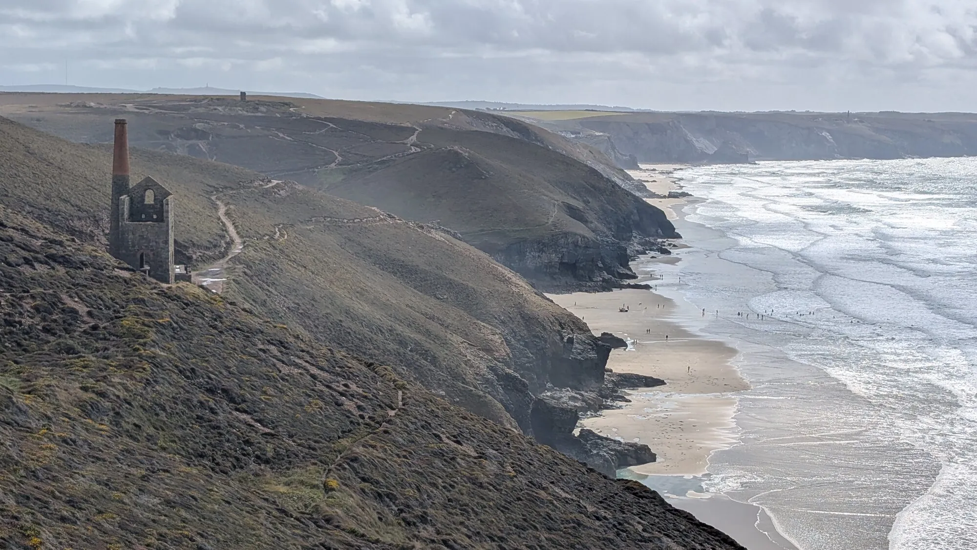 The Wheal Coates engine house stands on the cliff above Chapel Porth between St Agnes and Porthtowan on Cornwall's Tin Coast, with the surf-curled bay below and the wider mining landscape of cliffs and engine-house ruins beyond. The National Trust car park sits at the top, giving access to the South West Coast Path and the engine house itself.