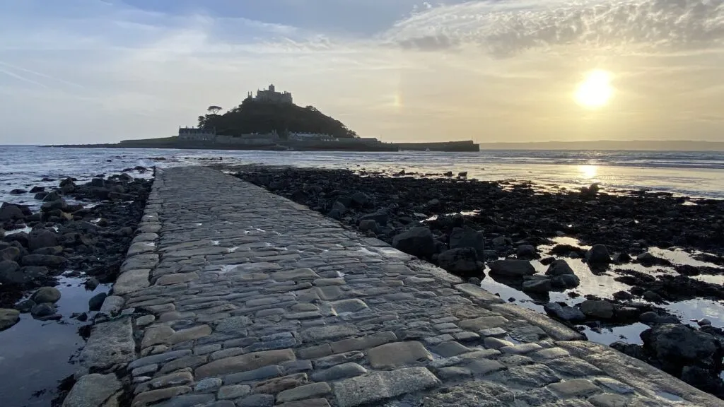 The cobbled causeway to St Michael's Mount in Marazion at low tide, the medieval castle silhouetted on its tidal island against a low sun, with kelp-strewn rocks either side of the granite path.