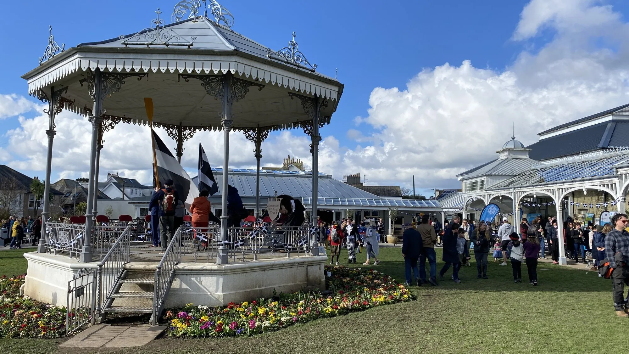 The Victorian wrought-iron bandstand in Gyllyngdune Gardens at the Princess Pavilion, Falmouth, with people on the bandstand holding Cornish St Piran flags during a community celebration, flower beds in the foreground and the conservatory of the Princess Pavilion visible to the right under a bright sky.