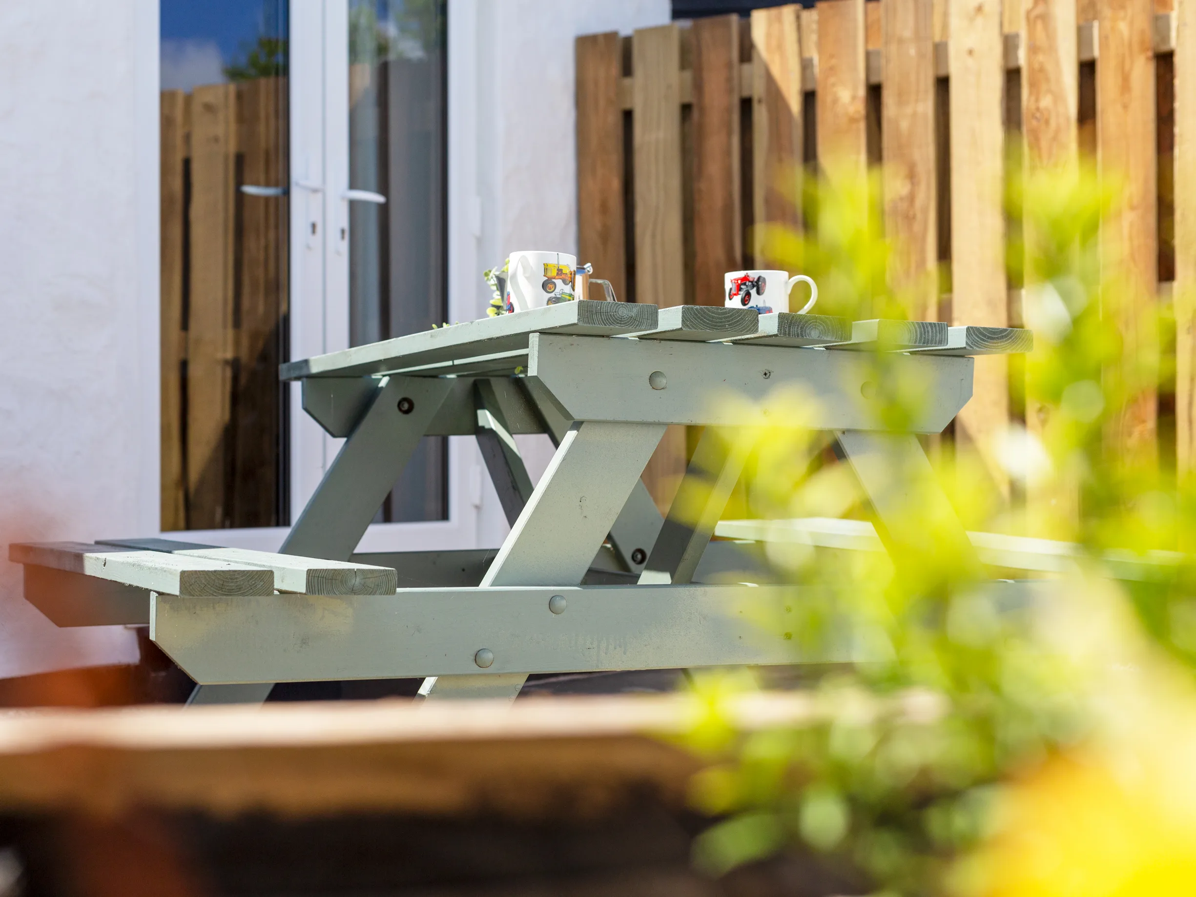 Two coffee mugs on a wooden bench on the patio outside the cottage, French doors open behind.