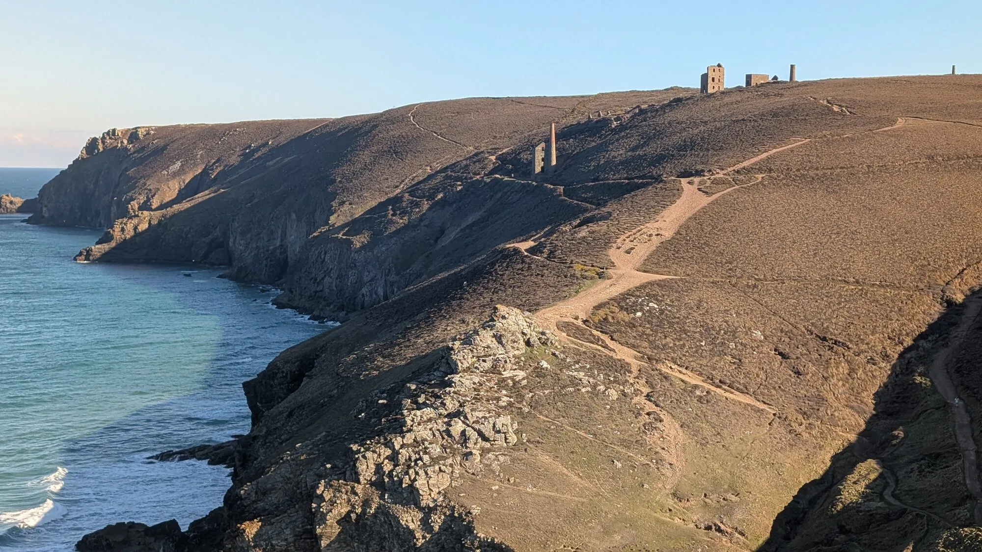 Late winter afternoon light on the Wheal Coates engine houses near St Agnes, with the ruined mining stack and engine house standing on the cliff top above turquoise sea, the dry brown vegetation of a Cornish winter all around.