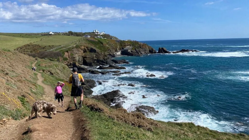 Walkers and a dog on the South West Coast Path with the Lizard headland and lighthouse in the distance, on a sunny day with rocky coves below the path.