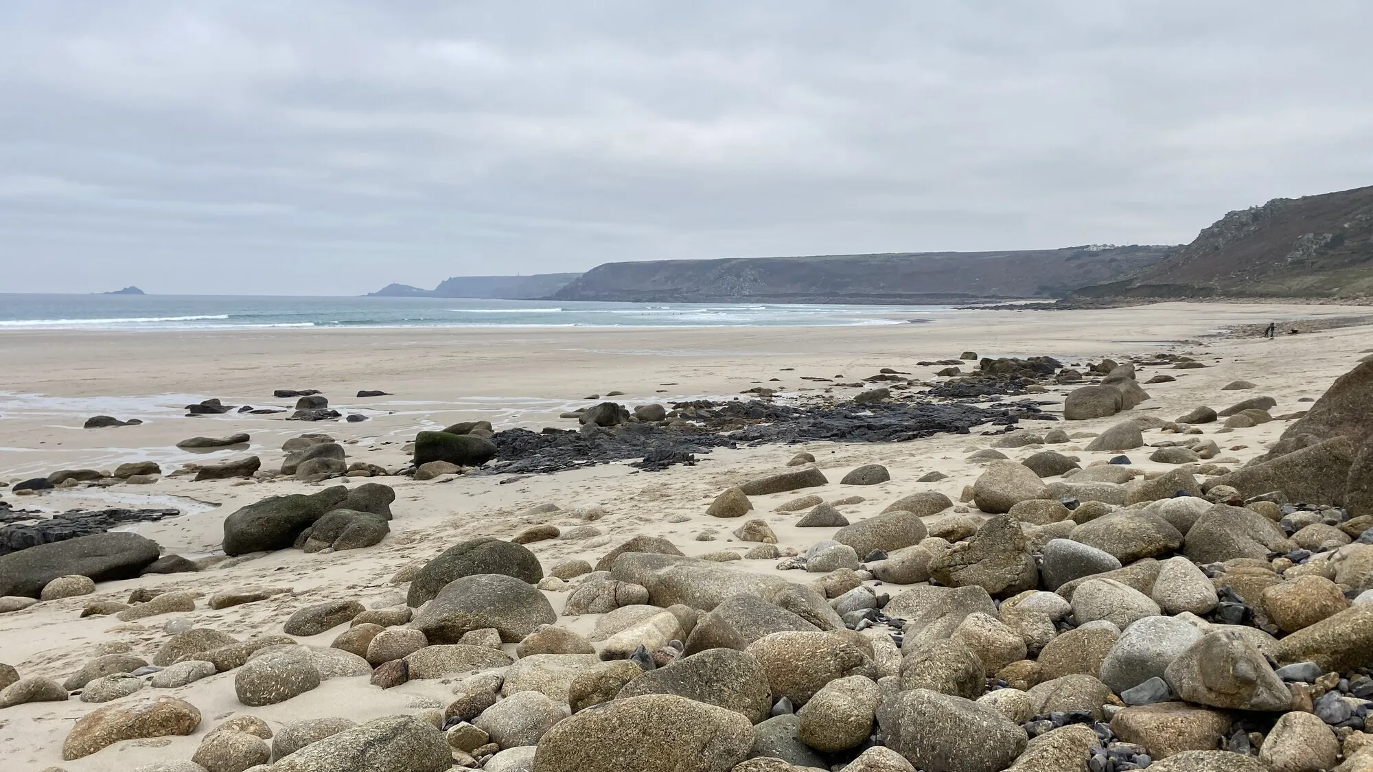 The wide sandy curve of Sennen Cove (Whitesand Bay) on Cornwall's far western coast, with rolling Atlantic surf, surfers in the lineup, and rugged headlands in the distance reaching towards Land's End under a clear summer sky.