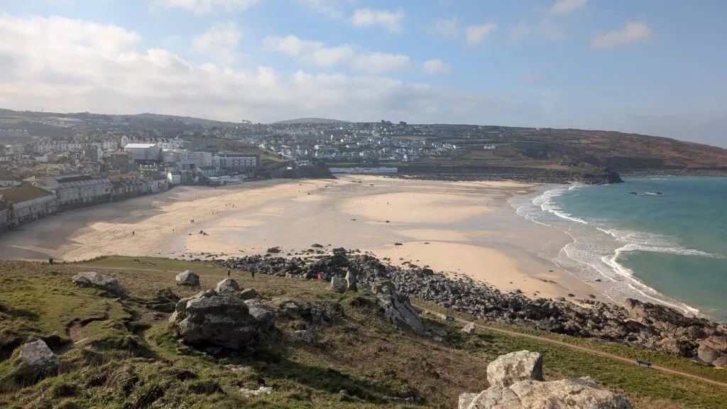 Porthmeor Beach in St Ives viewed from the headland called The Island, a wide arc of golden sand at low tide, the white-washed town and the Tate St Ives visible behind, a clear winter morning.