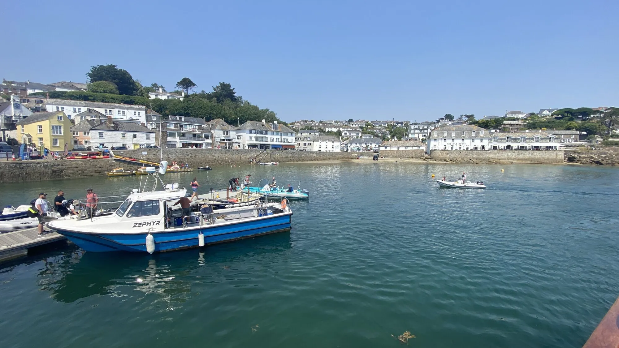 St Mawes harbour on a clear summer day: a blue and white passenger boat moored at the pontoon in the foreground, with the white-painted houses and inns of St Mawes village rising up the hillside behind under a deep blue sky.