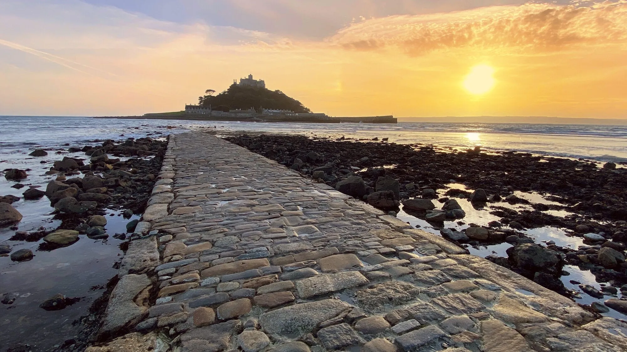 St Michael's Mount silhouetted against an orange sunset sky, with the cobbled granite causeway stretching across the seabed at low tide and kelp-strewn rocks and wet sand on either side.