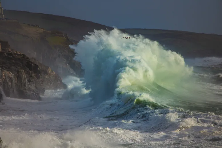 Huge winter storm waves crashing against the rugged Cornish coastline, sending sheets of white spray high into the grey sky.