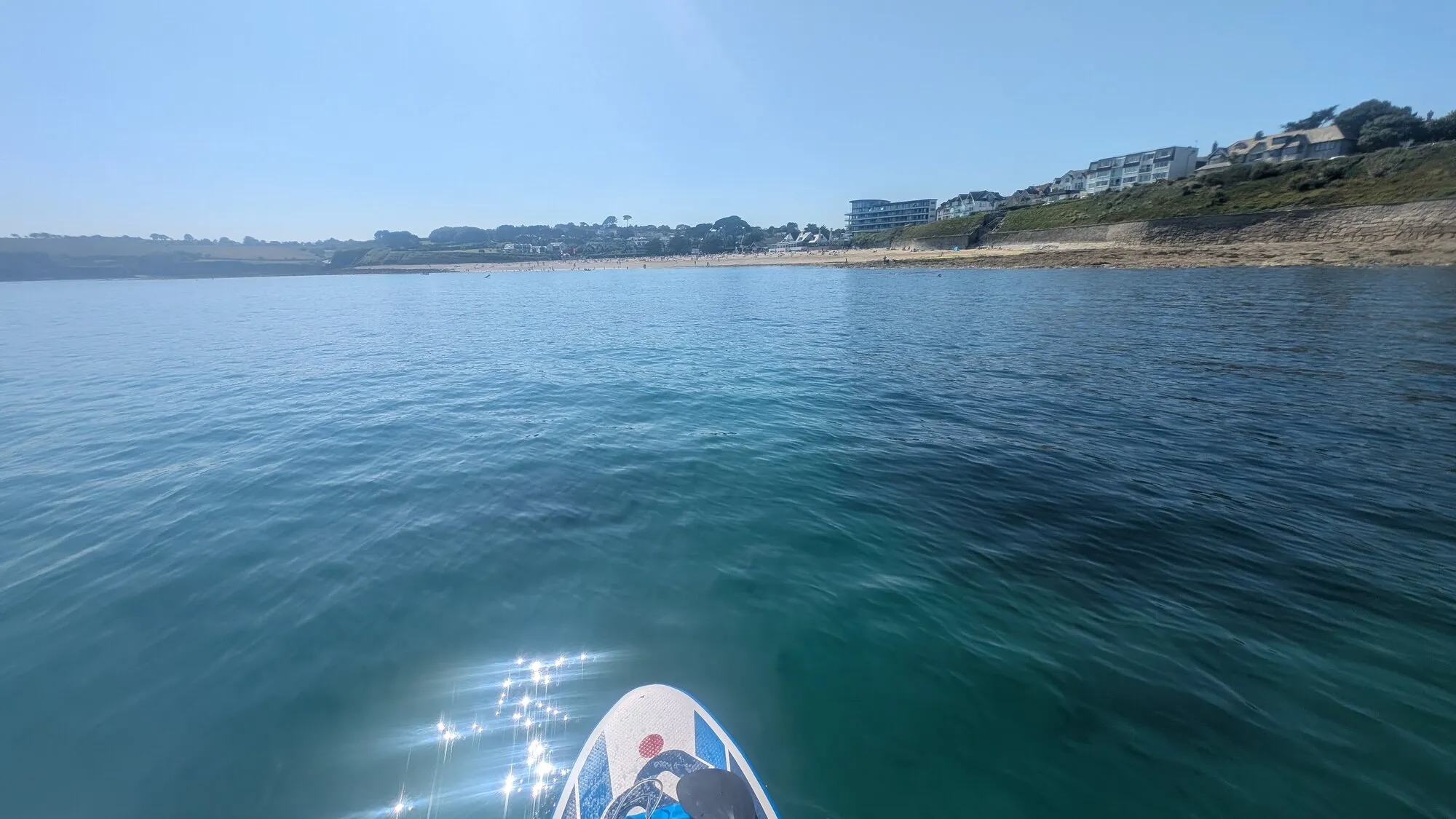 Looking back at Gyllyngvase Beach from a paddleboard on calm blue water, with the curve of golden sand and Falmouth's cliffside houses in the distance under a clear summer sky.