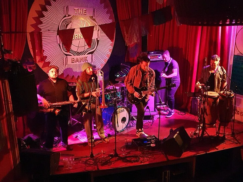 The Barefoot Bandit playing a live gig at the Cornish Bank in Falmouth: a five-piece band on stage under red lights, with saxophone, trombone, guitar, drums and percussion, performing in front of the venue's distinctive Cornish Bank backdrop.