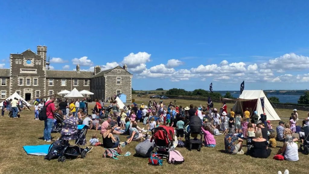Families gathered on the grass for a summer event at Pendennis Castle in Falmouth, with the historic granite garrison buildings and a glimpse of the sea behind them.