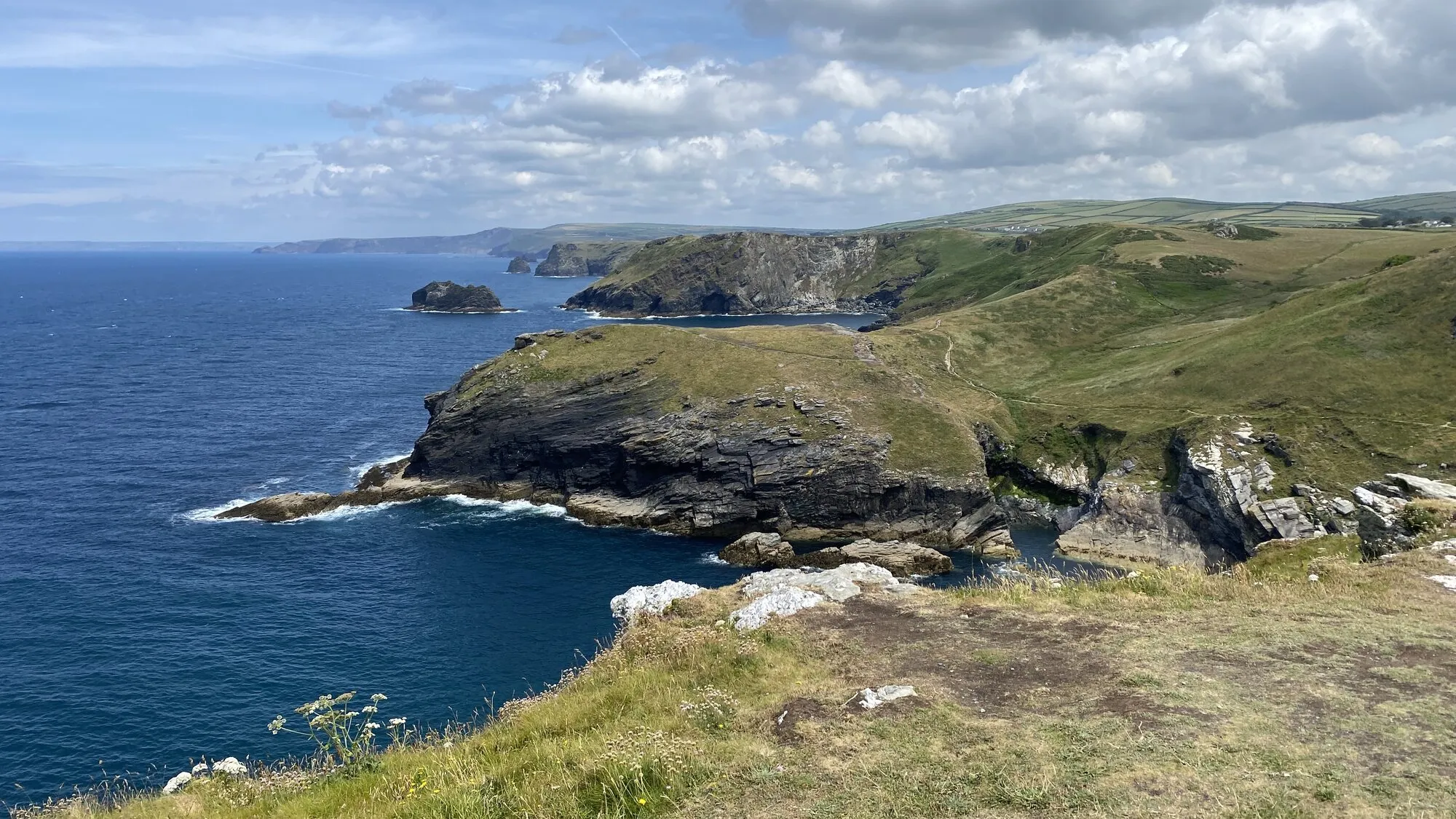 The North Cornwall coast looking north from above Tintagel, with dark slate cliffs dropping to a deep blue sea, a small rocky island offshore, and the green clifftop path of the South West Coast Path curving away along the headland towards Boscastle under a bright cloud-scattered sky.