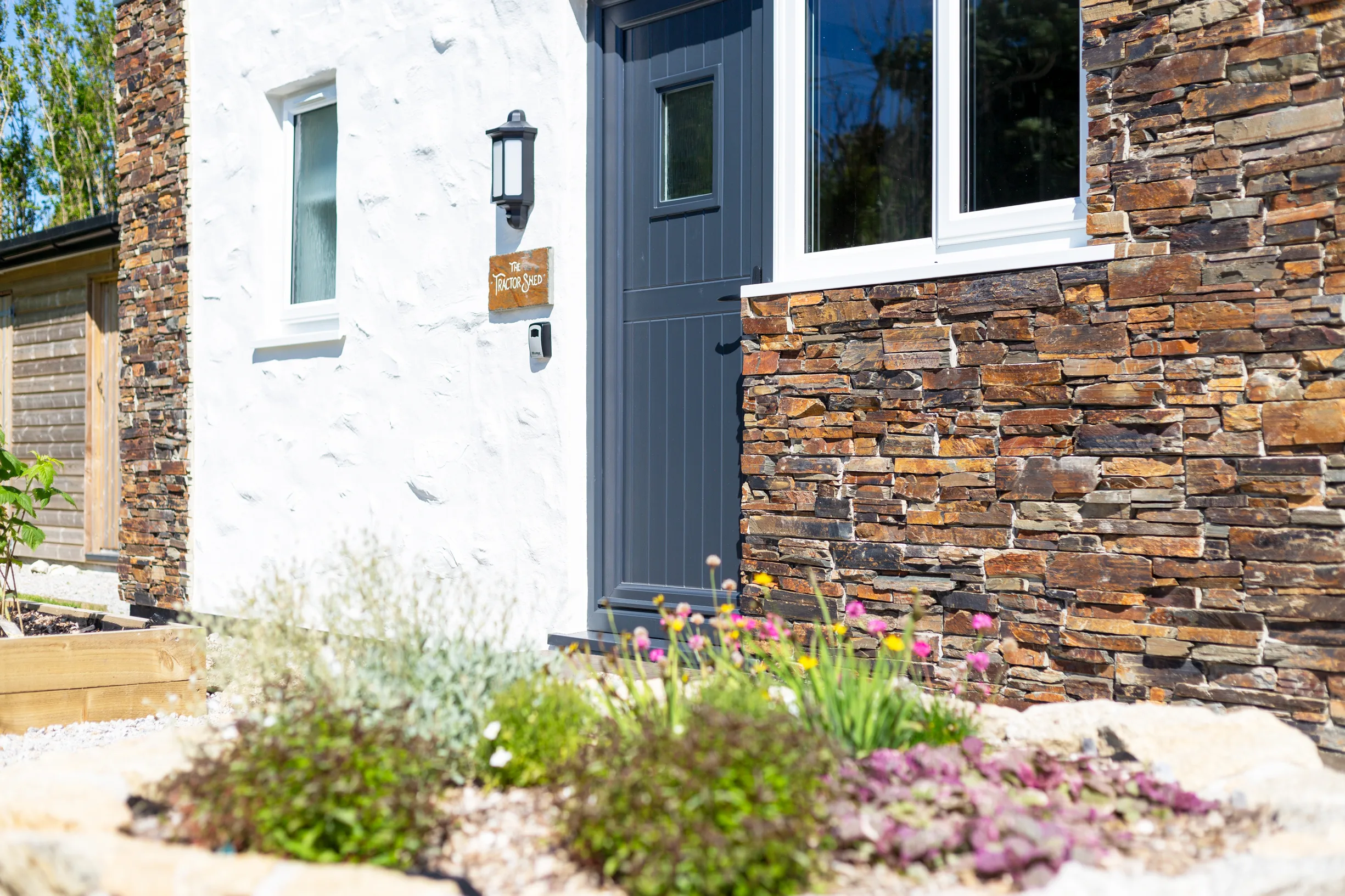 The Tractor Shed exterior. Slate stone wall, dark blue door, white render, garden in the foreground.