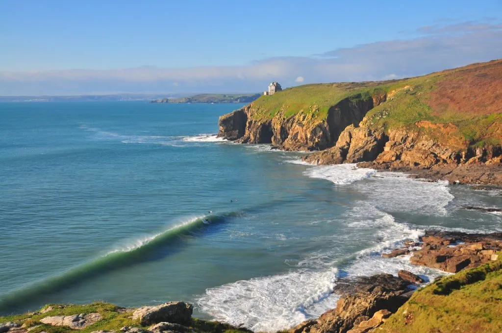 A long peeling wave wraps around a Cornish headland into a sandy cove with surfers in the line-up and a white coastguard lookout on the cliff above, on a bright sunny day.