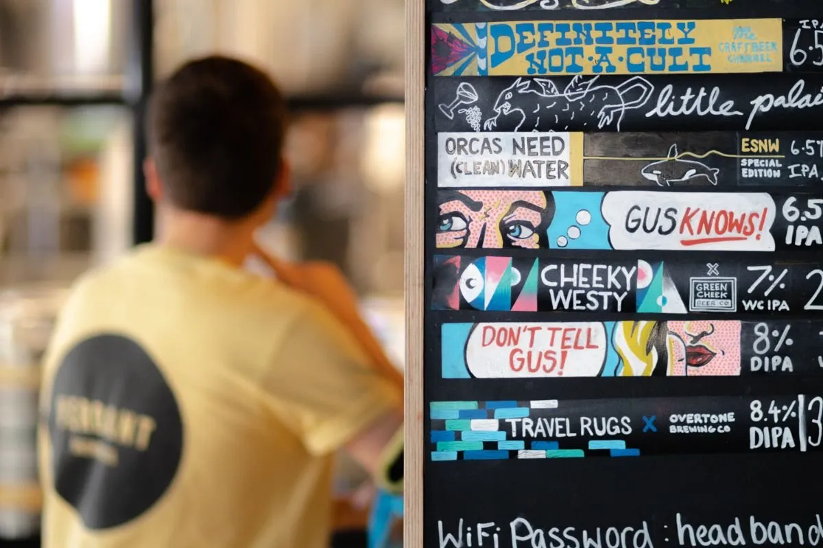 Inside the Verdant Brewery taproom in Penryn: a hand-painted chalkboard menu listing the day's beers (Definitely Not A Cult, Little Palais, Orcas Need Clean Water, Gus Knows, Cheeky Westy, Don't Tell Gus, Travel Rugs) with prices and ABVs in bright colours, a member of the brewery staff in a yellow Verdant t-shirt walking past in soft focus to the left, and the working brewery visible behind them through the open partition between the taproom and the brewery floor.