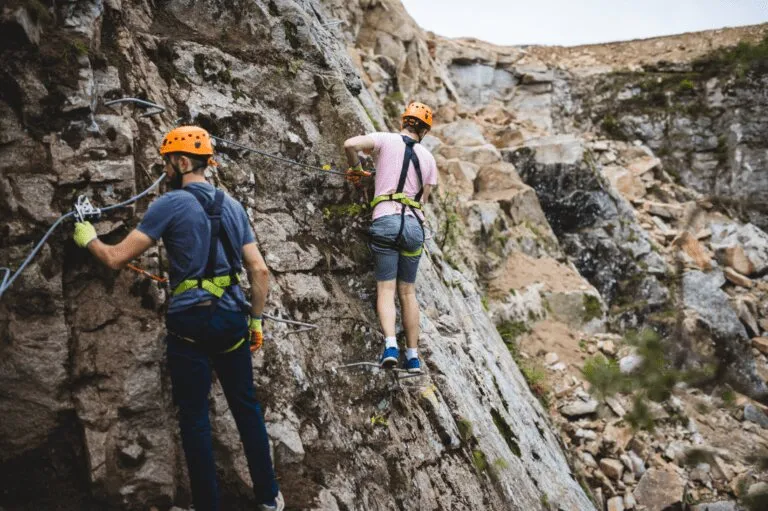 Two climbers in orange helmets and harnesses traverse the granite rock face of a former Cornish quarry on the Via Ferrata Cornwall iron stairway.