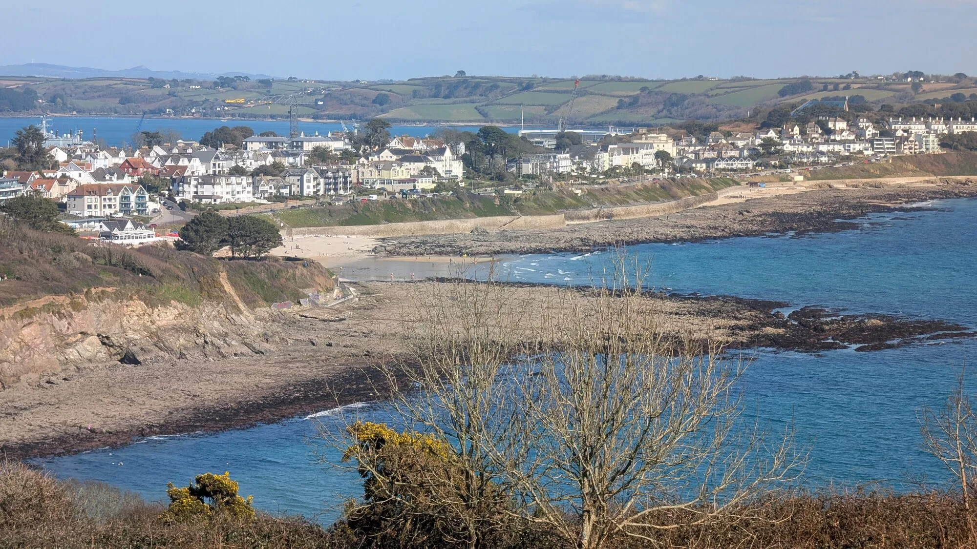 An autumn view across Falmouth's cliffside hotels, Castle Beach and Gyllyngvase from the headland, with bare branches in the foreground and the calm waters of Falmouth Bay leading away to the distant hills.