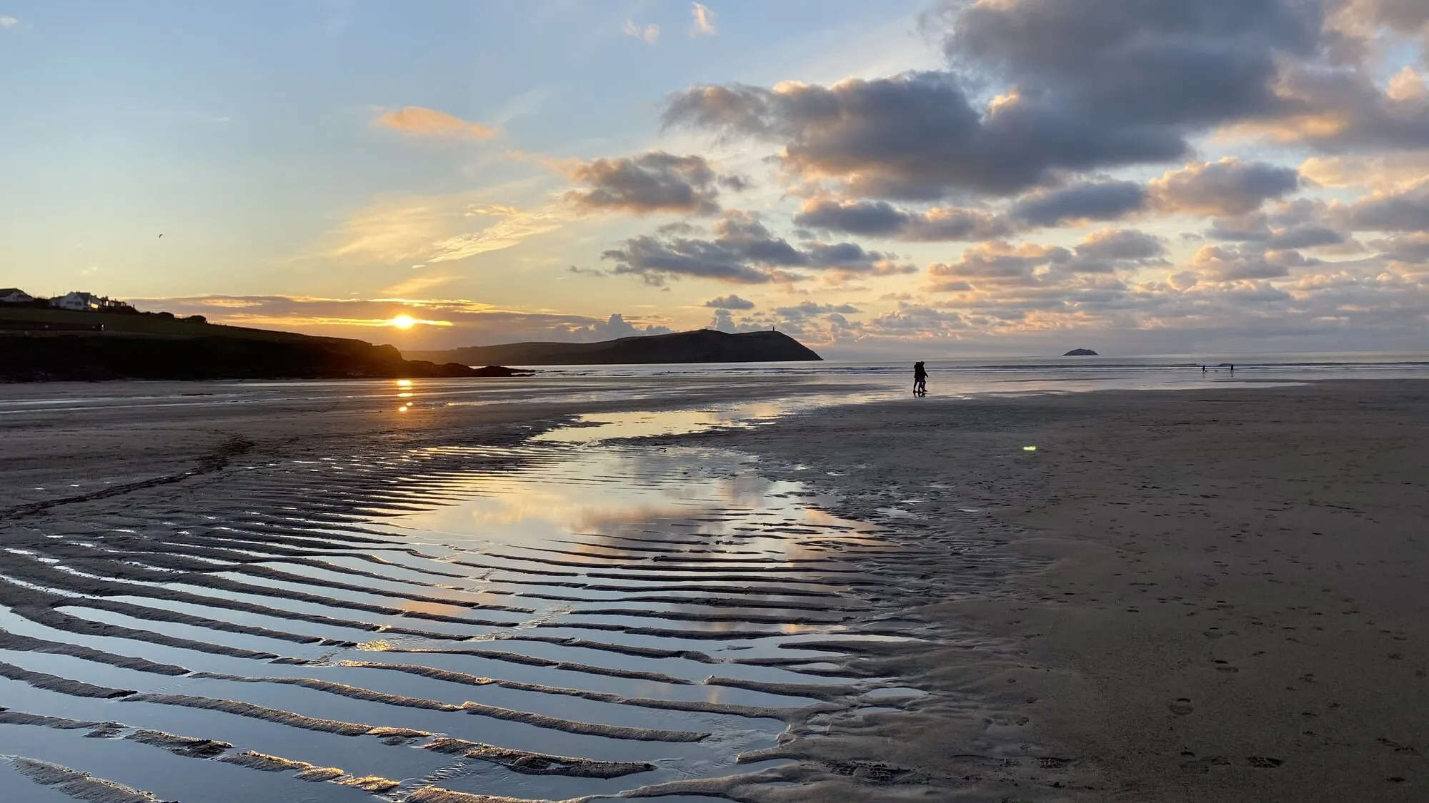 A couple in silhouette walking an empty Cornish beach at low tide, with the sun setting behind a distant headland and rippled wet sand reflecting the pastel sky.